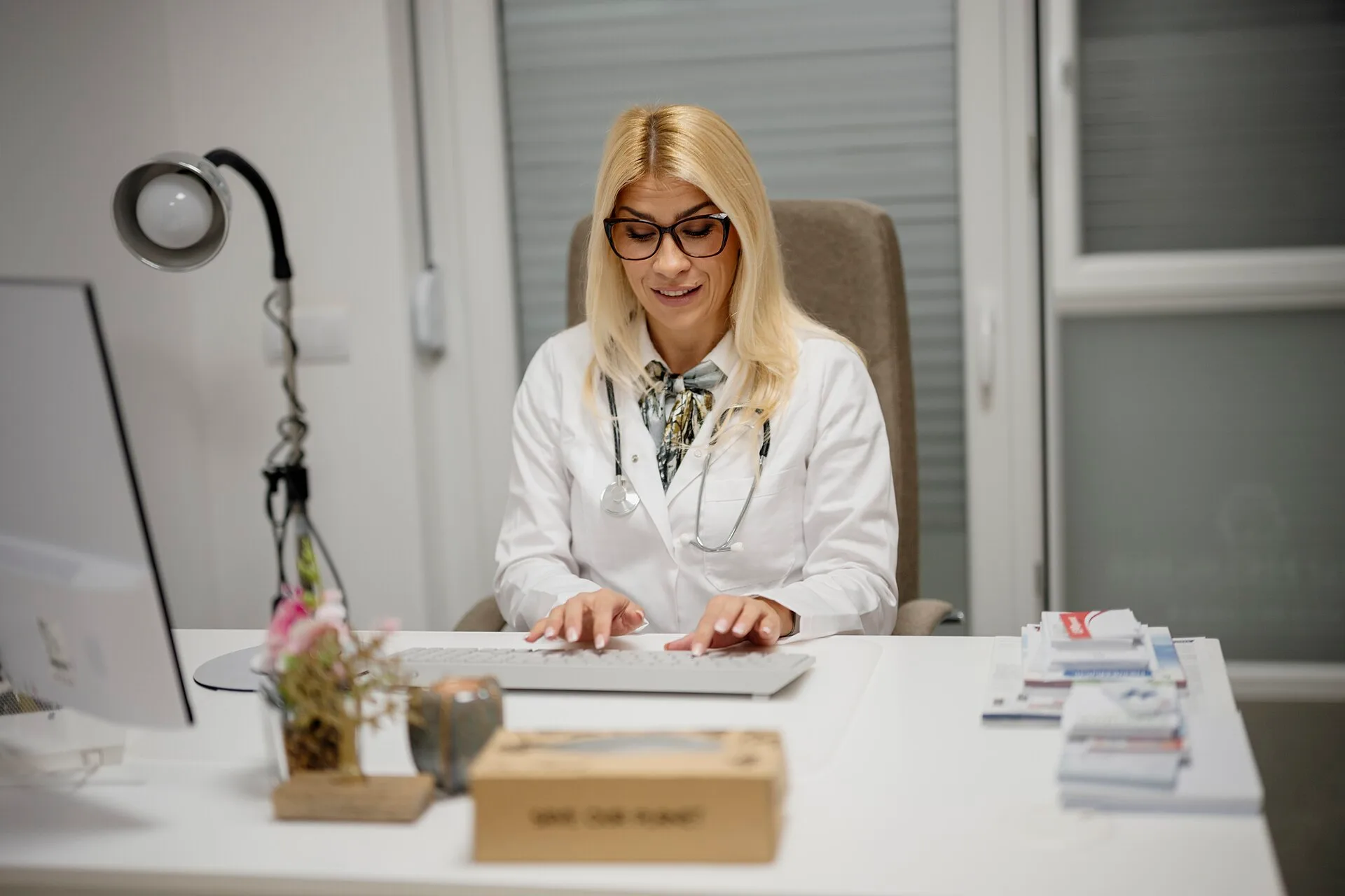 A doctor sits at her desk in a medical office. She is wearing a white coat and glasses. She uses a computer and takes notes while looking engaged. The Healthcare SEO Best Practices Worth Adopting First