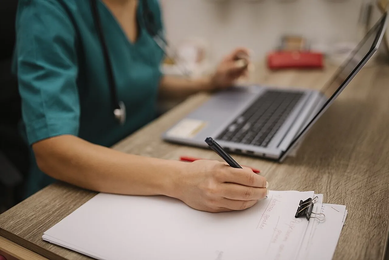 A healthcare professional in scrubs writes notes beside an open laptop at a desk.