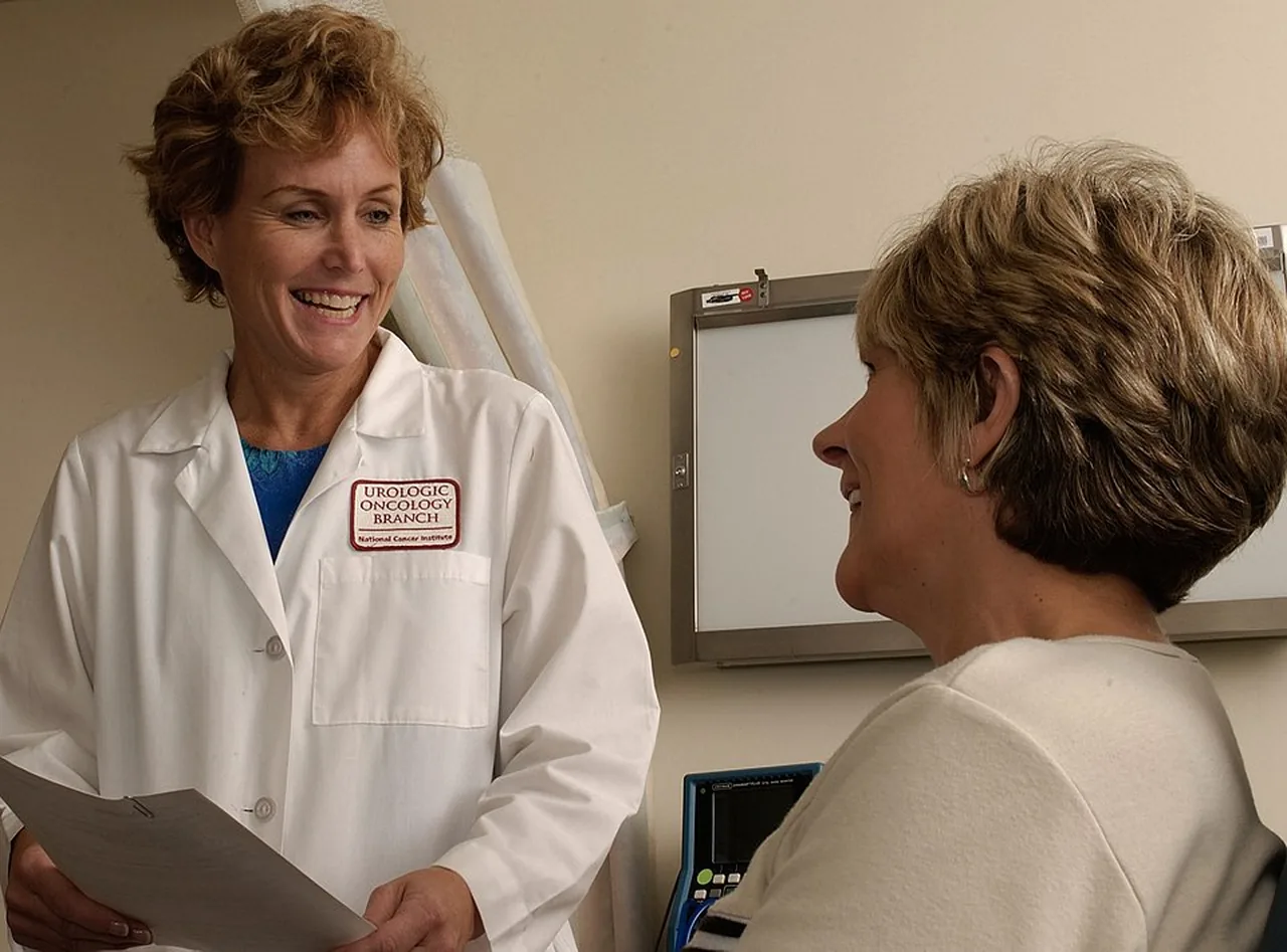 A physician in a white coat speaks with a seated patient in a clinical examination room.
