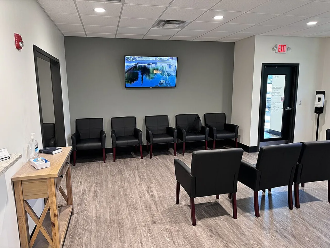 An empty medical waiting room has black chairs, a television, and an exit door.